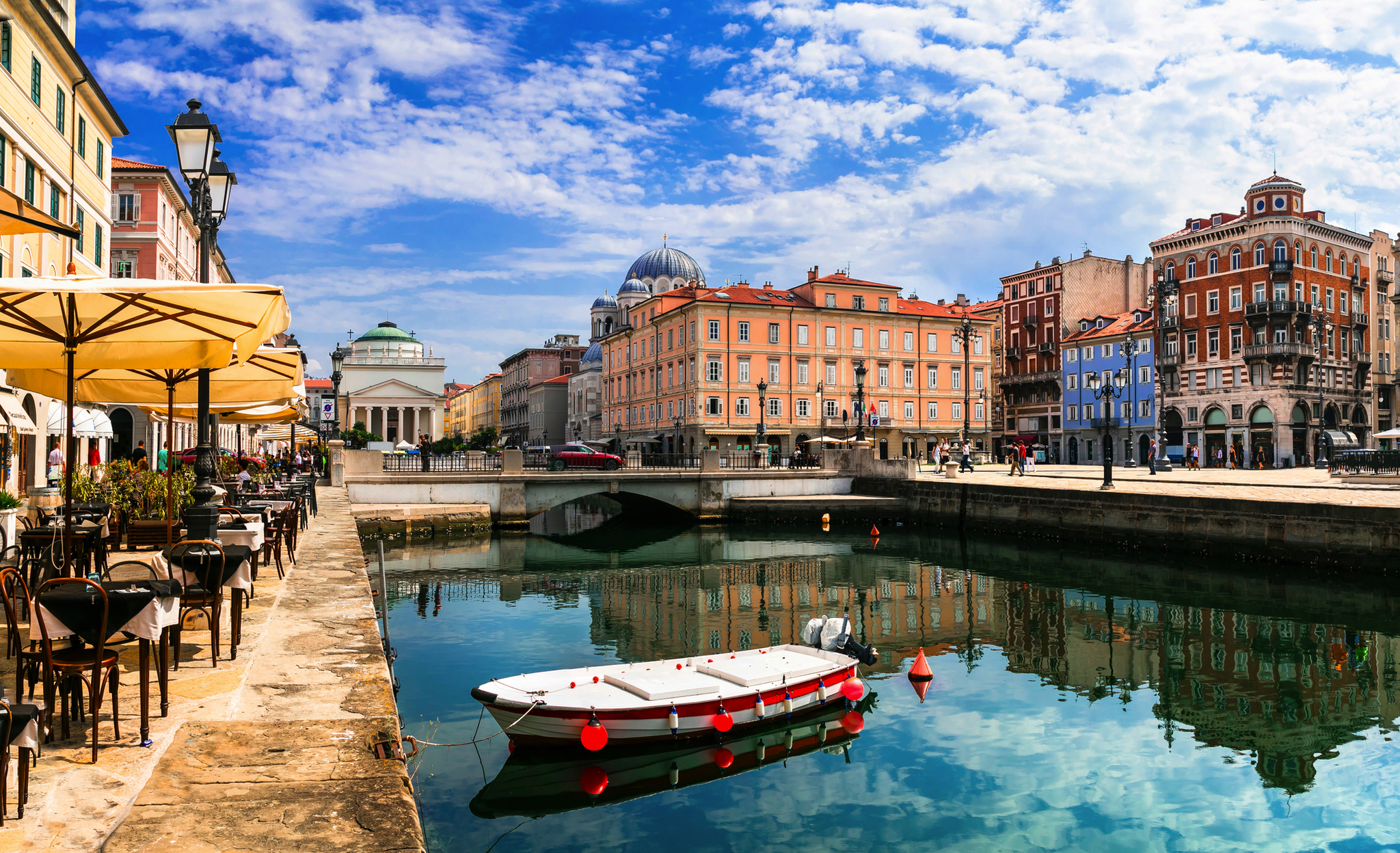 Triest Kanal mit Boot, umgeben von historischen Gebäuden und Straßencafés unter blauem Himmel.